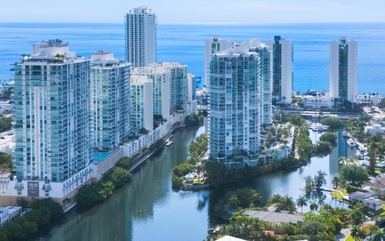 Aerial photograph of Sunny Isles Beach from the bay side: condominium towers side by side along a waterway, floor above floor, with the ocean behind them.